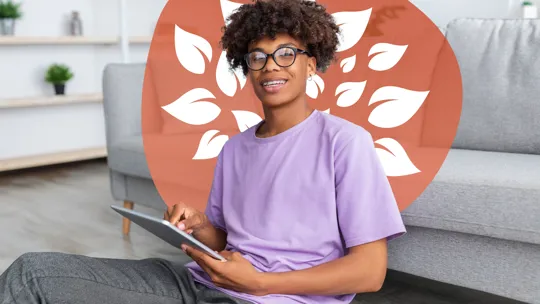 A teenager with glasses and braces, smiling and looking at the camera while holding a tablet, sitting on the floor in a casual setting.