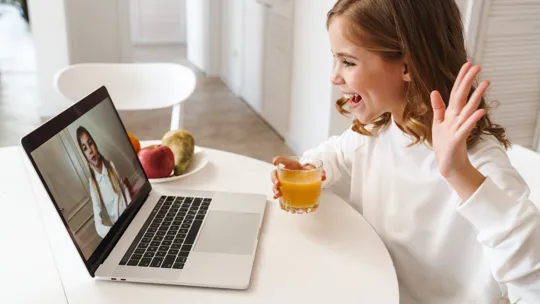Smiling girl waving at a laptop screen during a video call, with a glass of juice in hand.