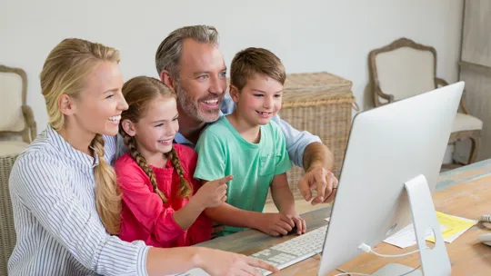 Family gathered around a computer screen, smiling and engaged in online learning