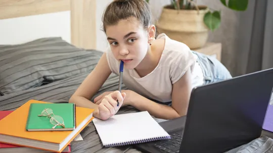 Teen girl lying on bed, thoughtfully holding a pen while studying with laptop and folders.