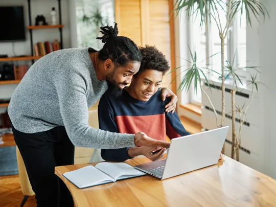 Father smiling and guiding his teenage son through online studies on a laptop at home.