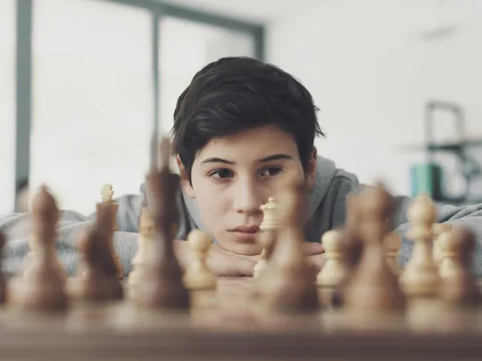 Focused boy resting his head on folded arms, observing a chessboard closely.