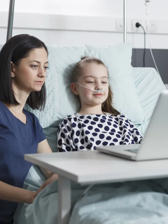 Girl recovering in hospital using a laptop with supportive adult by her side