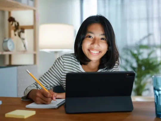 A homeschooling female student with short black hair studying at her bedroom desk, smiling at the camera.