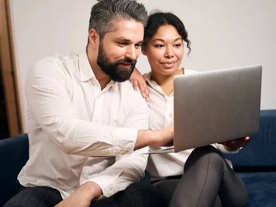 Couple seated closely on a sofa, attentively viewing a laptop screen in a relaxed home setting.