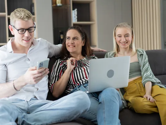 Three people sitting on a sofa smiling while looking at a laptop and phone together