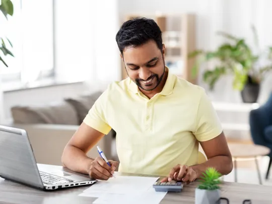 Man smiling while working on a laptop and with a calculator, possibly managing homeschool finances for his family in Riyadh.