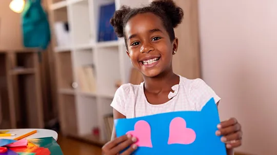 A homeschooling young girl smiling holding up a creative craft picture with pink hearts on blue card.