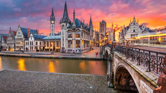 Historic buildings along a canal in Ghent at sunset with glowing streetlights and reflections