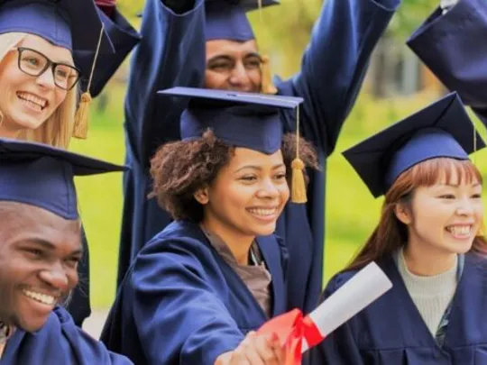 A group of university graduates in blue gowns and mortarboards smiling to the right and holding heir degree scrolls.
