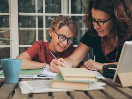 A parent assists a smiling child with their homeschool studies at a table with books and a laptop, illustrating personalized education options for families homeschooling in Riyadh.