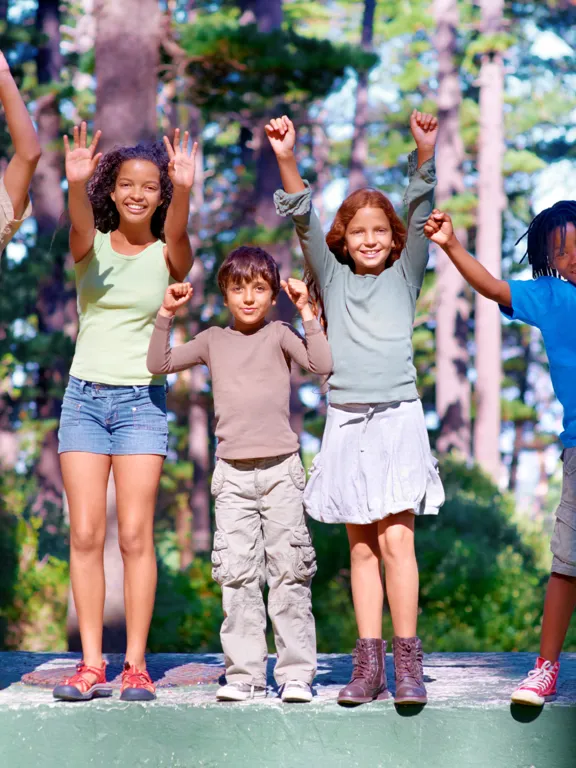 Five children standing with arms raised, smiling in a wooded outdoor setting.