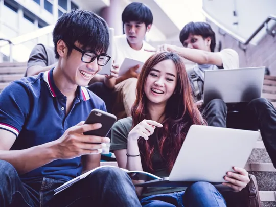Modern homeschooling approaches in Penang, showing students using a mix of traditional notebooks and digital devices like laptops and phones for collaborative study on steps.