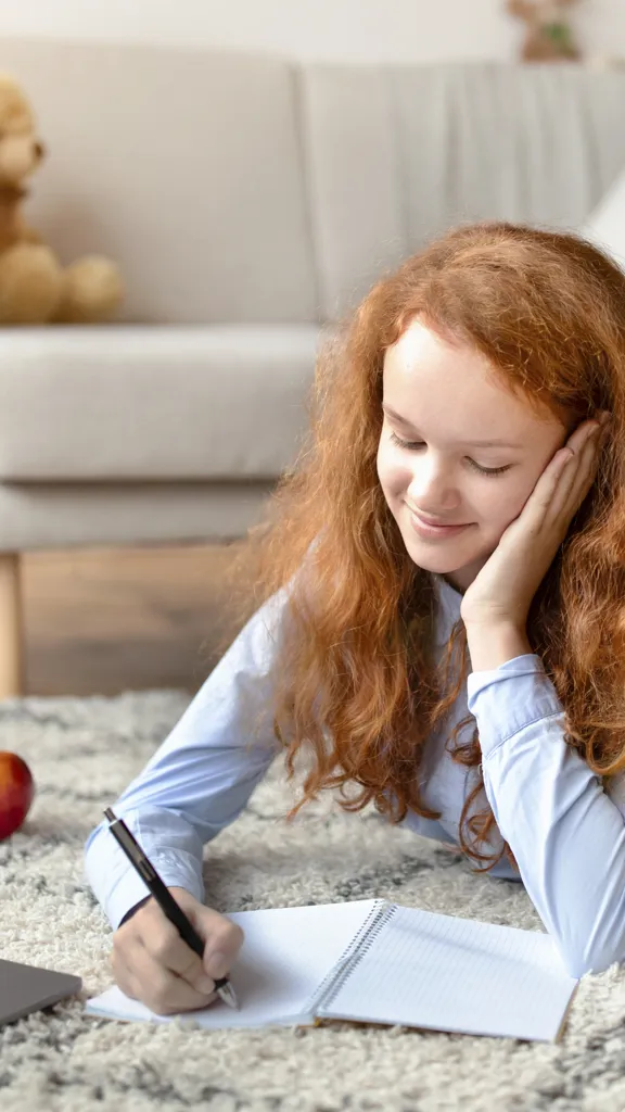 Smiling girl lying on rug, writing in a notebook with a laptop and apple nearby.