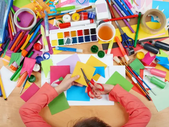 Overhead view of a child using scissors for a colourful paper craft activity surrounded by art supplies