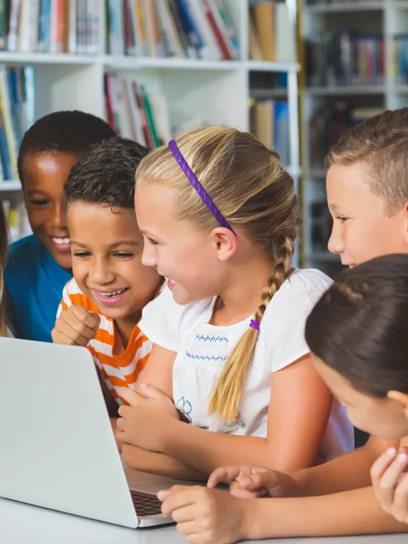 Group of smiling primary-aged children gathered around a laptop in a school library