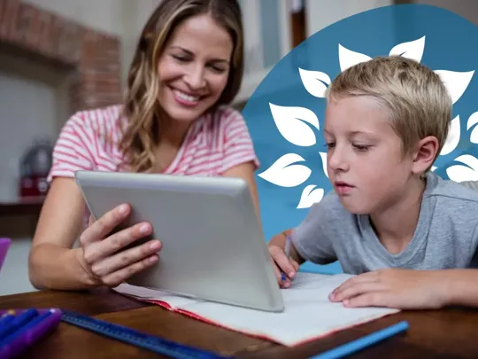 A homeschooling mum and son sitting at a desk. The mum is smiling and showing her son something on a tablet device, which the boy is studying intently.