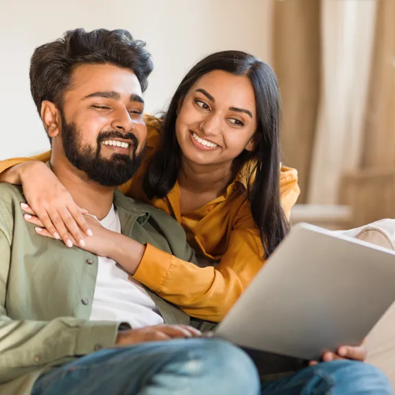 Couple smiling warmly while looking at a laptop together on a cosy living room sofa.