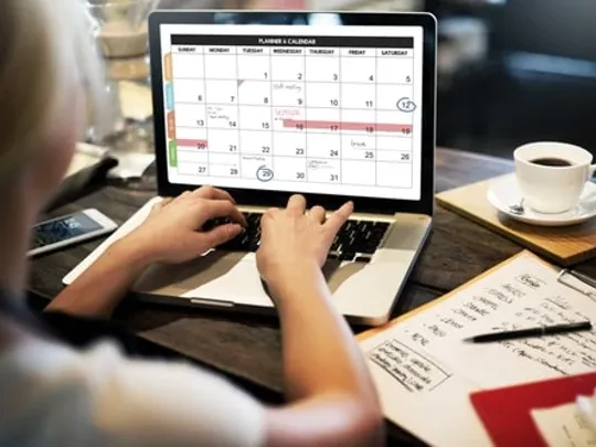 A photograph of a female homeschooling student with her back to the camera working on a laptop, which is displaying a calendar schedule. The student's paper assignment and pen are on the desk next to the laptop.