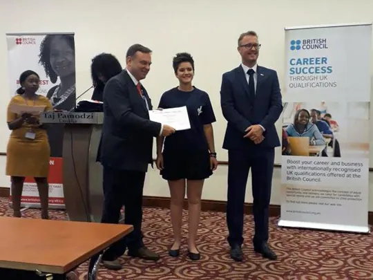 Wolsey Hall Oxford student Celia smiles at the camera and stands with two men whilst being awarded Outstanding Cambridge Learner Award.