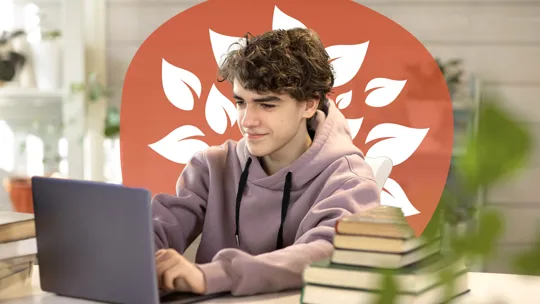 A young student with curly hair, wearing a hoodie, smiling slightly while working on a laptop at a desk with stacked books.