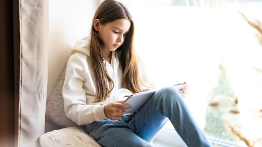 Young girl with long hair reading on a tablet while sitting on a cushion by a bright window.