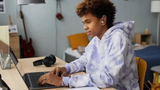 Focused boy wearing a hearing aid, studying at his desk with a laptop and notebook.
