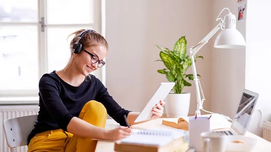 An older female student working at her desk with her laptop and reading notes she is making.