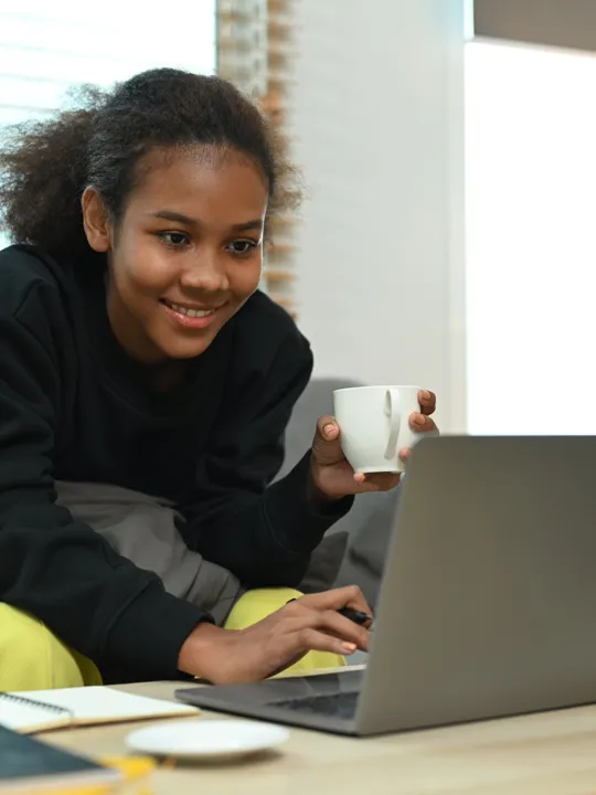 Smiling student with a mug in hand using a laptop at home, seated on a sofa with notebooks on the table.