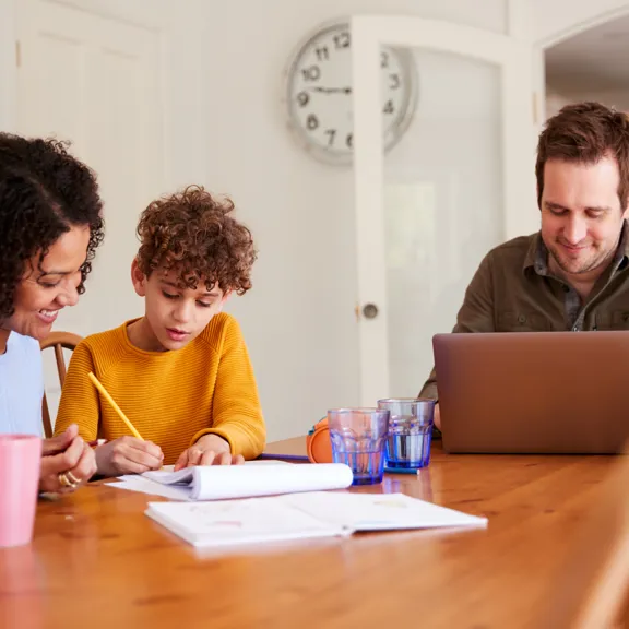 Mother helping child with work at a table while father works on a laptop nearby