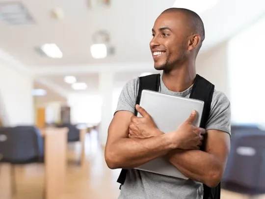 A young man with a black rucksack smiling to the left, holding a laptop to his chest with both arms.