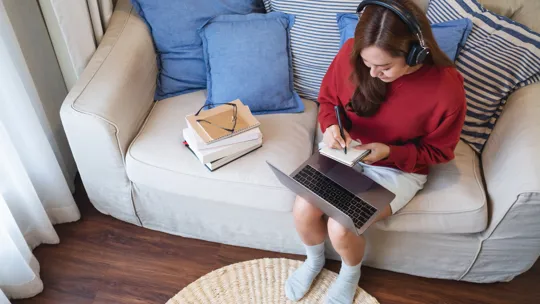 Teenage girl writing notes with laptop on lap and headphones on, sitting on a sofa with study materials beside her