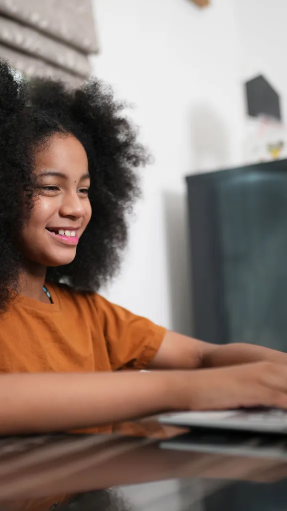 Young girl smiling as she types on a laptop at a kitchen table.