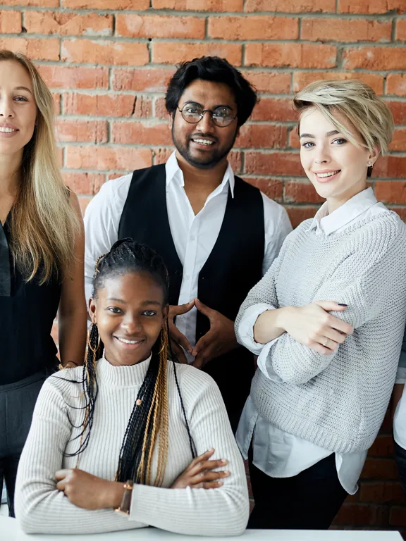 Group of five young professionals smiling together in a modern office with brick walls.