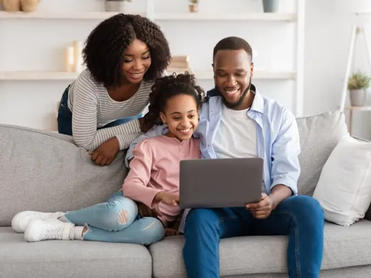 A homeschooling mum, dad and young daughter in their living room smiling at a laptop. The dad and daughter are sat on the sofa, whilst the mother is leaning on the back of it.