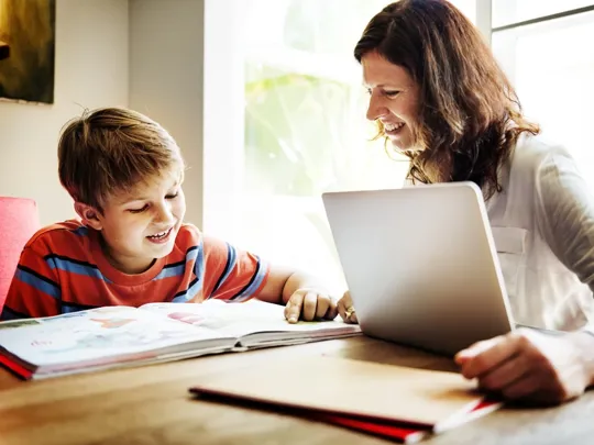 A mum helping her young son homeschool. They are both sat smiling at a desk with a laptop and a textbook.