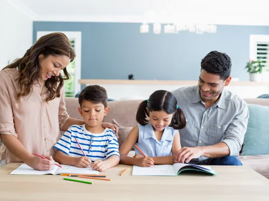 Two parents supporting their young children with schoolwork around a living room table.