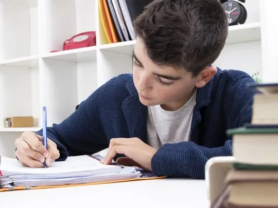 A homeschooling young boy with short black hair writing in a notepad at a desk with books.